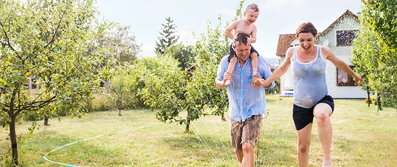 Familie med mor og far samt barn på farens skuldre hopper igennem en vandspreder i haven en sommerdag. Moren er gravid. I baggrunden ses familiens hus.